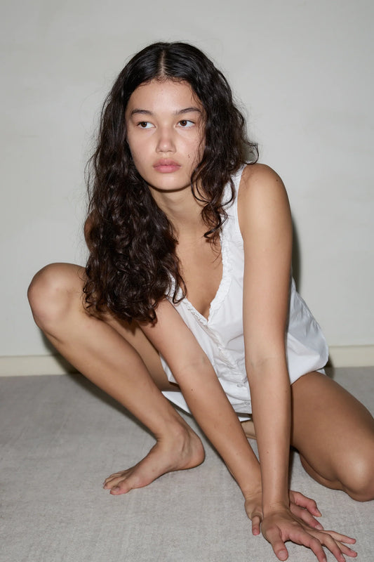 A woman with long, wavy dark hair wears a sleeveless white PJ Short Set, crouching on the floor and gazing to the side with a neutral expression against a plain background.
