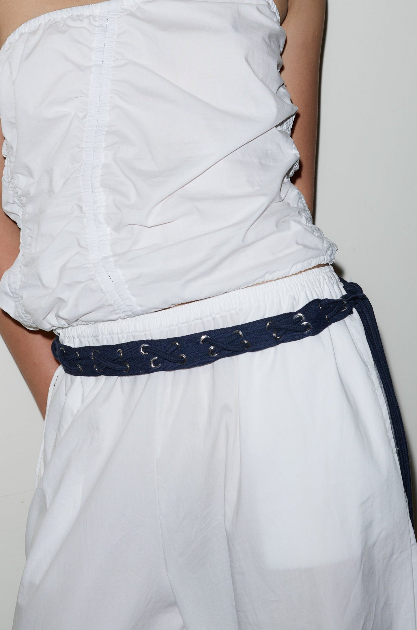 A person stands against a white background wearing a white sleeveless ruched top, loose white pants, and the Eyelet Belt - Navy.