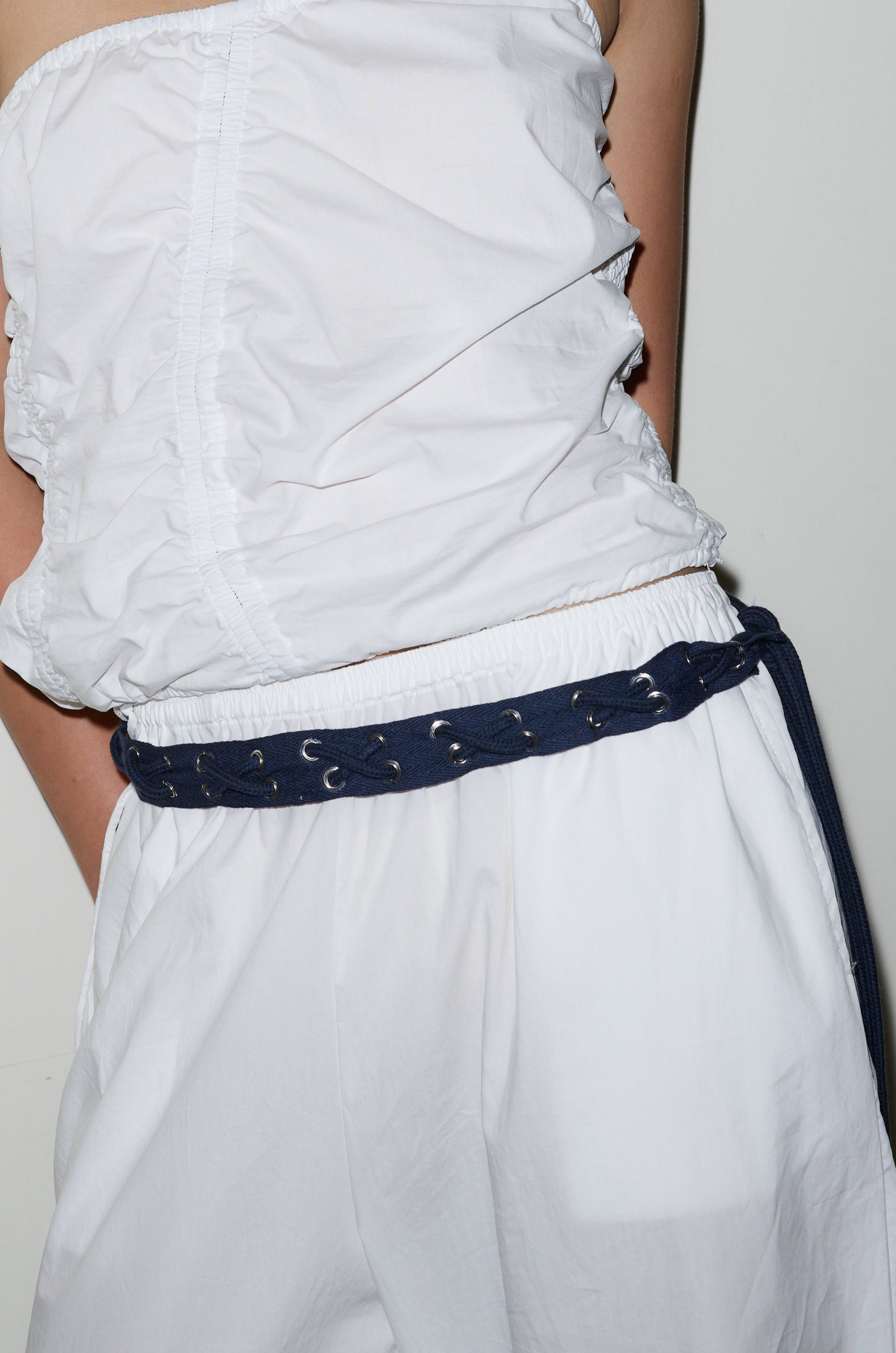 A person stands against a white background wearing a white sleeveless ruched top, loose white pants, and the Eyelet Belt - Navy.