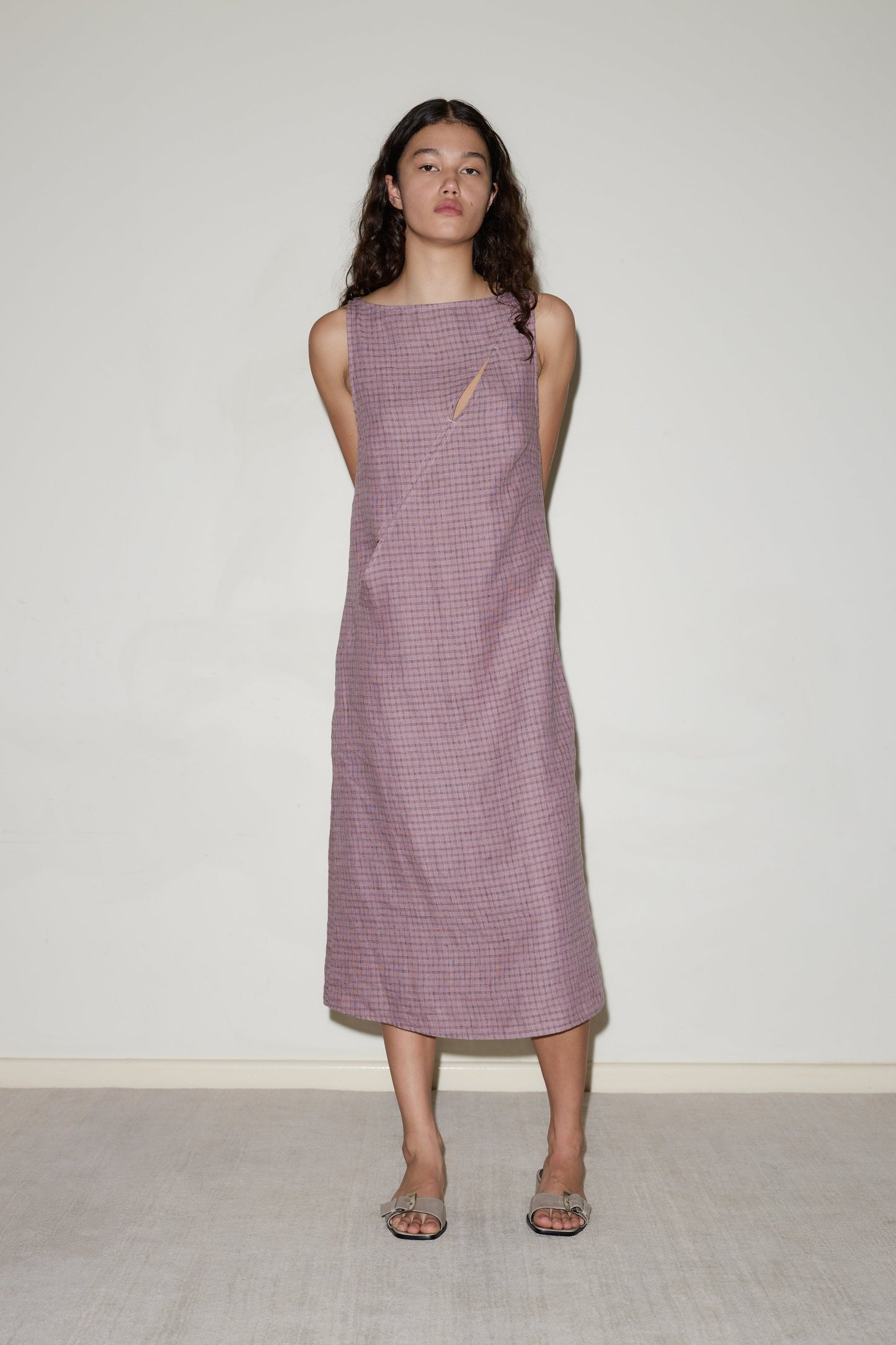 A woman with long, wavy brown hair stands against a light wall, wearing the sleeveless, loose-fit High Tie Dress in textured lavender with a small neckline cutout. She has her arms behind her back and is wearing sandals.