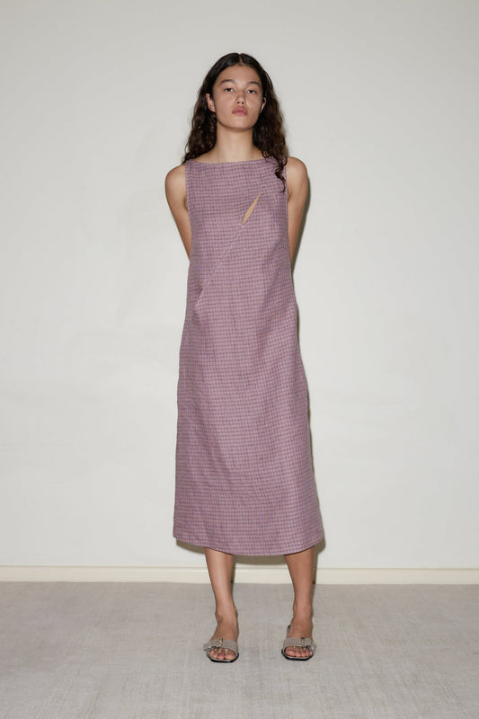 A woman with long, wavy brown hair stands against a light wall, wearing the sleeveless, loose-fit High Tie Dress in textured lavender with a small neckline cutout. She has her arms behind her back and is wearing sandals.