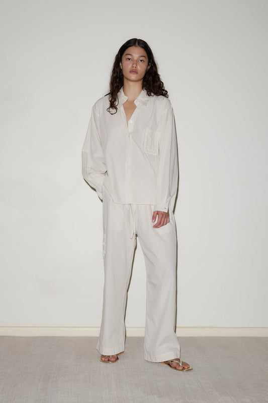 A person stands against a plain white wall, barefoot on a light-colored floor, wearing the Cotton Pyjama Set: a loose-fitting, off-white button-up shirt with front pocket and matching wide-leg pants. They have long, wavy hair.