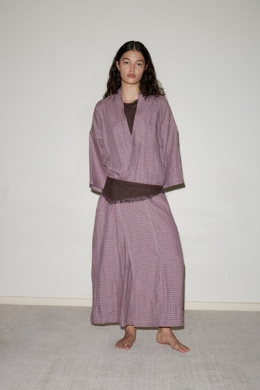 A woman with long, wavy hair stands barefoot against a plain wall, wearing the 02 Robe—a loose-fitting, mauve kimono-style top and wide-legged skirt with a subtle pattern—layered over a dark shirt.