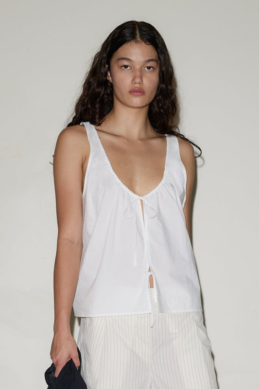 A woman with long, wavy dark hair wears the Cotton Bind Top— a sleeveless white V-neck style with a front tie—paired with light vertically striped pants, standing against a plain light background.