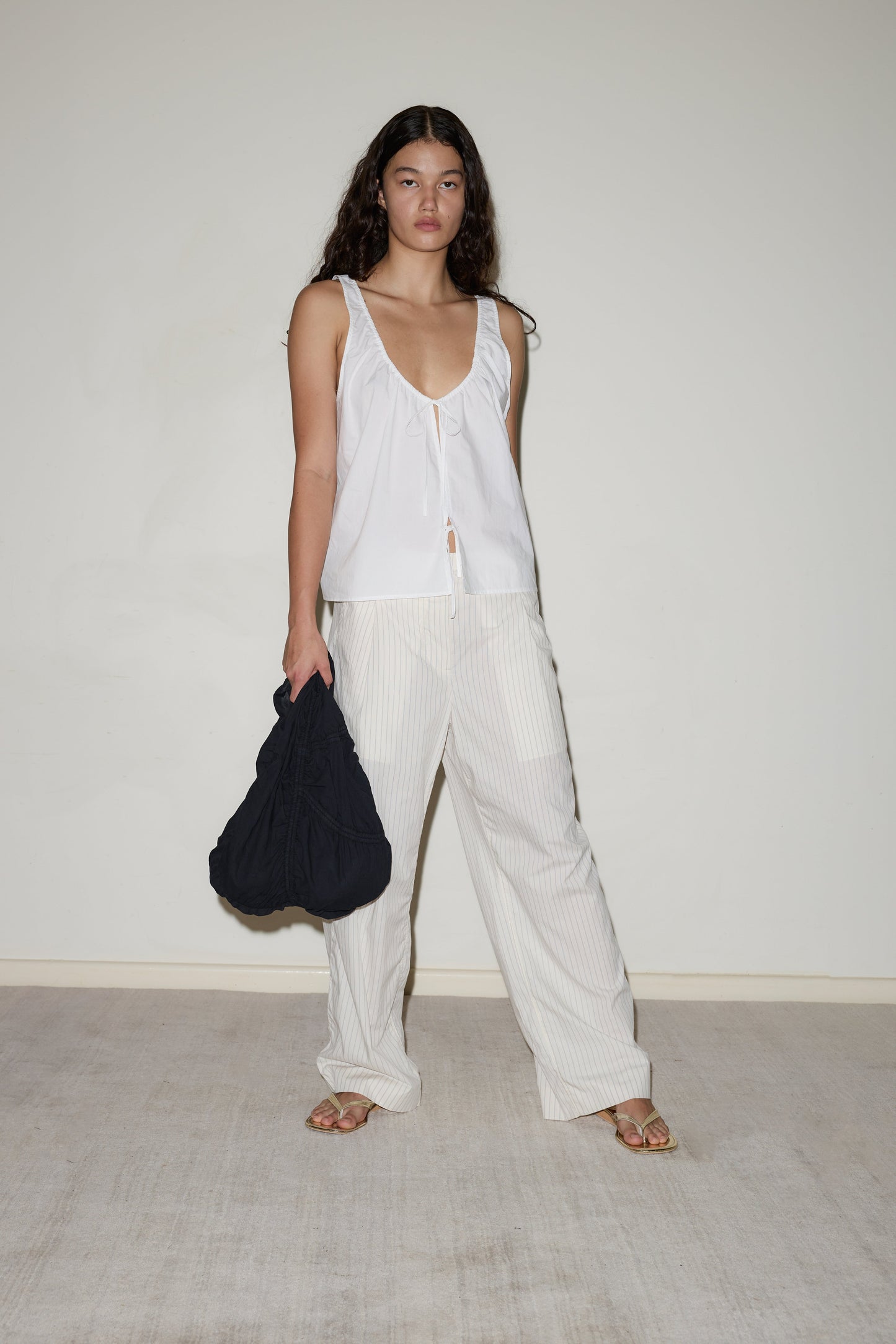 A woman with long curly hair wears light striped Twin Button Pants, a loose white sleeveless top, sandals, and holds a large black bag while standing against a plain light-colored wall.