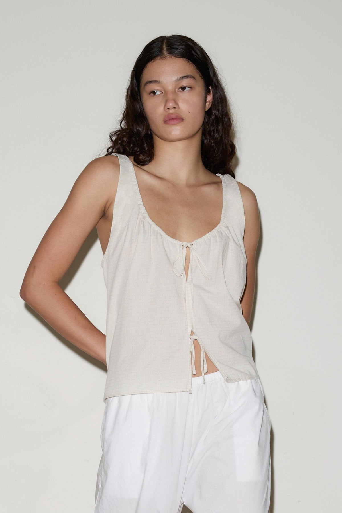 A woman with long, wavy dark hair wears the sleeveless Cotton Bind Top in off-white with a front tie and loose white pants, standing against a plain light-colored background.