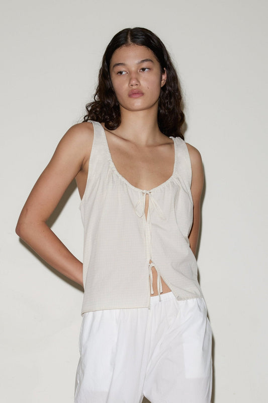A woman with long, wavy dark hair wears the sleeveless Cotton Bind Top in off-white with a front tie and loose white pants, standing against a plain light-colored background.