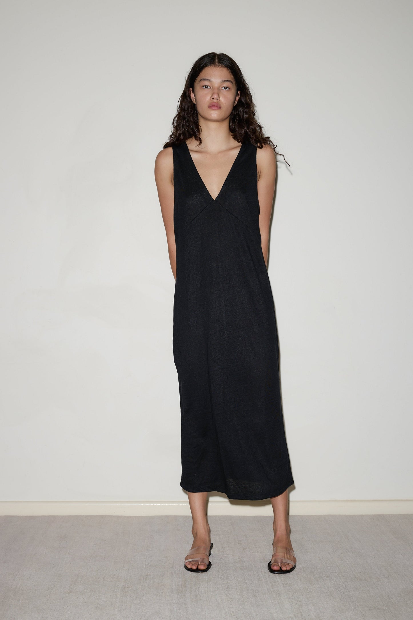 A woman with long curly hair stands against a plain light wall, wearing the Soft Tank Dress—a sleeveless black midi dress with a deep V-neck—paired with clear heeled sandals. She looks directly at the camera, arms behind her back.