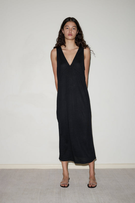 A woman with long curly hair stands against a plain light wall, wearing the Soft Tank Dress—a sleeveless black midi dress with a deep V-neck—paired with clear heeled sandals. She looks directly at the camera, arms behind her back.
