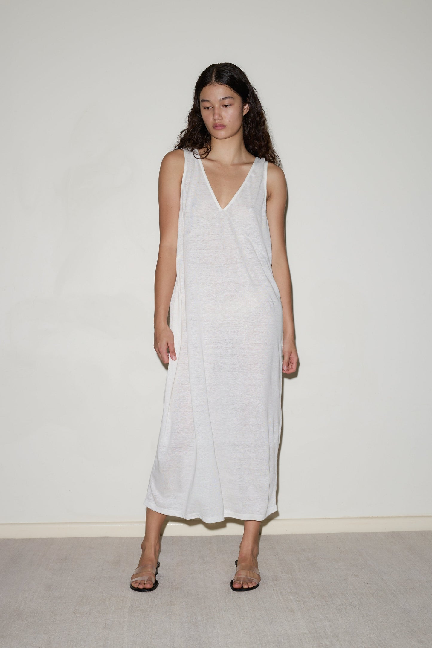 A woman with long dark hair stands against a plain light wall, wearing the Soft Tank Dress—a loose, sleeveless, semi-sheer white piece—paired with open-toed sandals. She gazes slightly downward with a neutral expression.