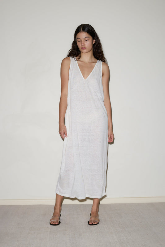 A woman with long dark hair stands against a plain light wall, wearing the Soft Tank Dress—a loose, sleeveless, semi-sheer white piece—paired with open-toed sandals. She gazes slightly downward with a neutral expression.