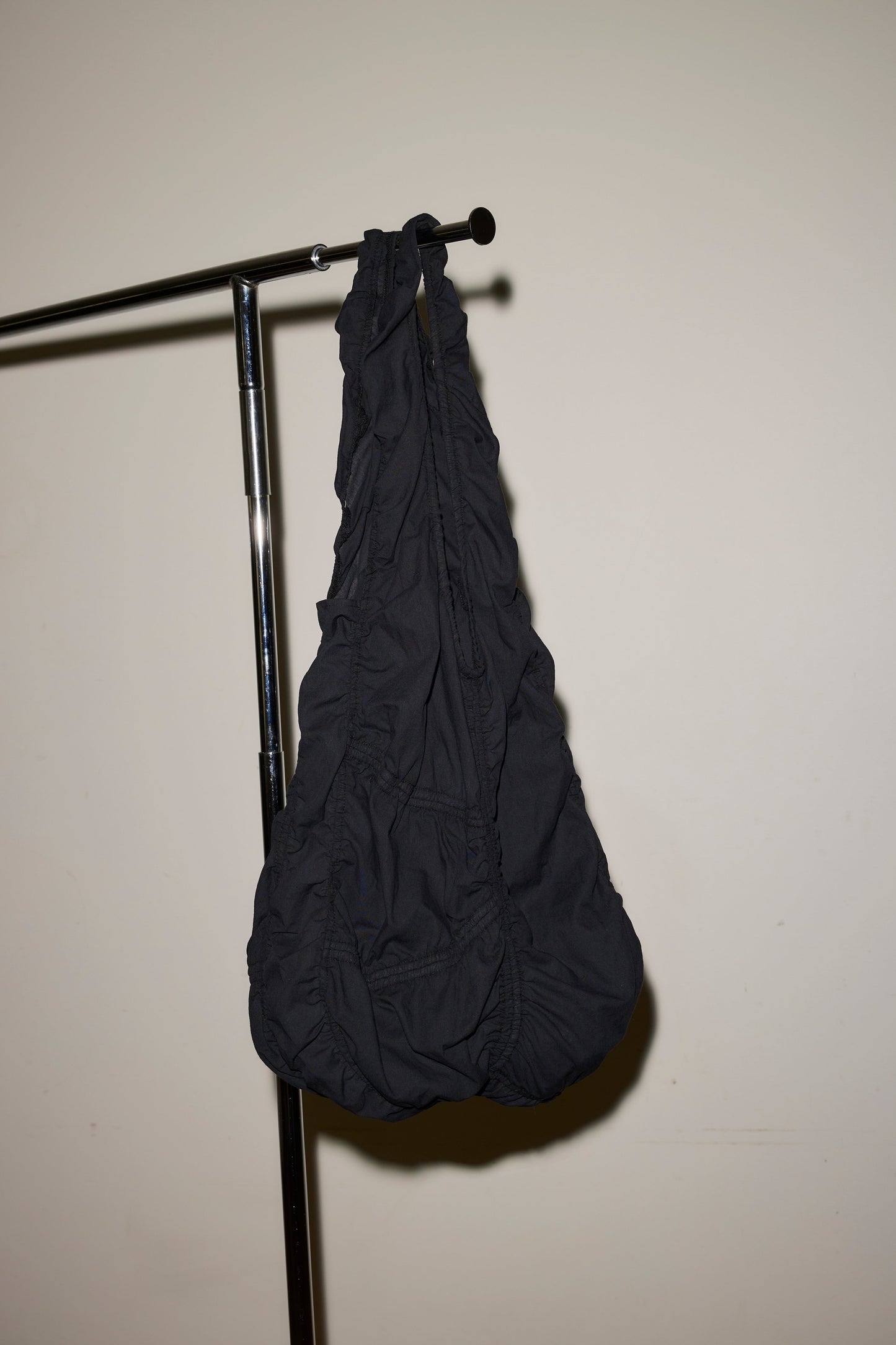 The Parachute Bag, crafted from black ruched fabric, hangs from a metal clothing rack set against a plain off-white wall.