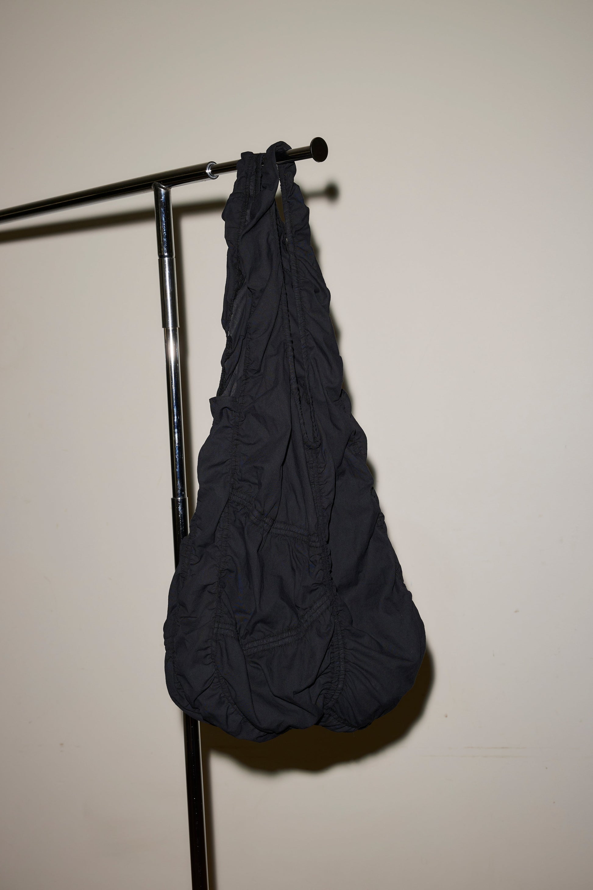 The Parachute Bag, crafted from black ruched fabric, hangs from a metal clothing rack set against a plain off-white wall.