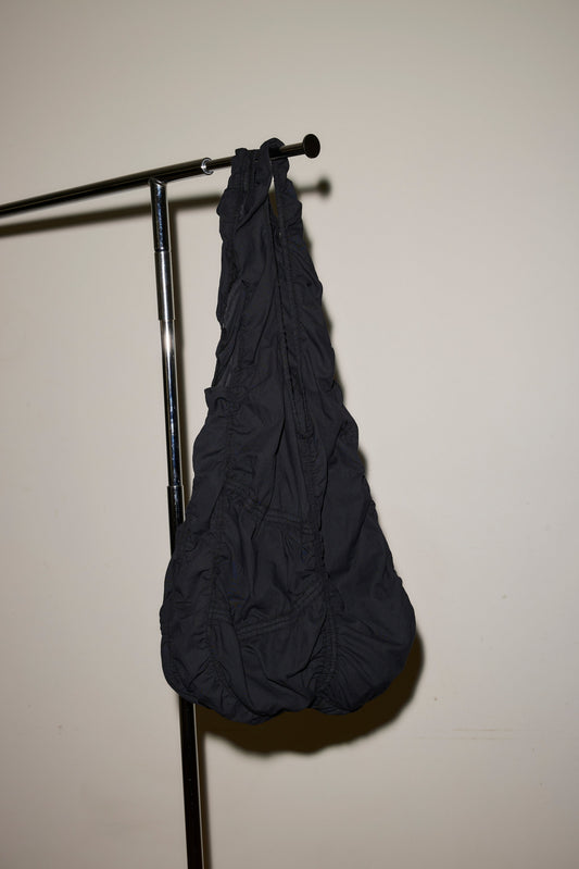 The Parachute Bag, crafted from black ruched fabric, hangs from a metal clothing rack set against a plain off-white wall.