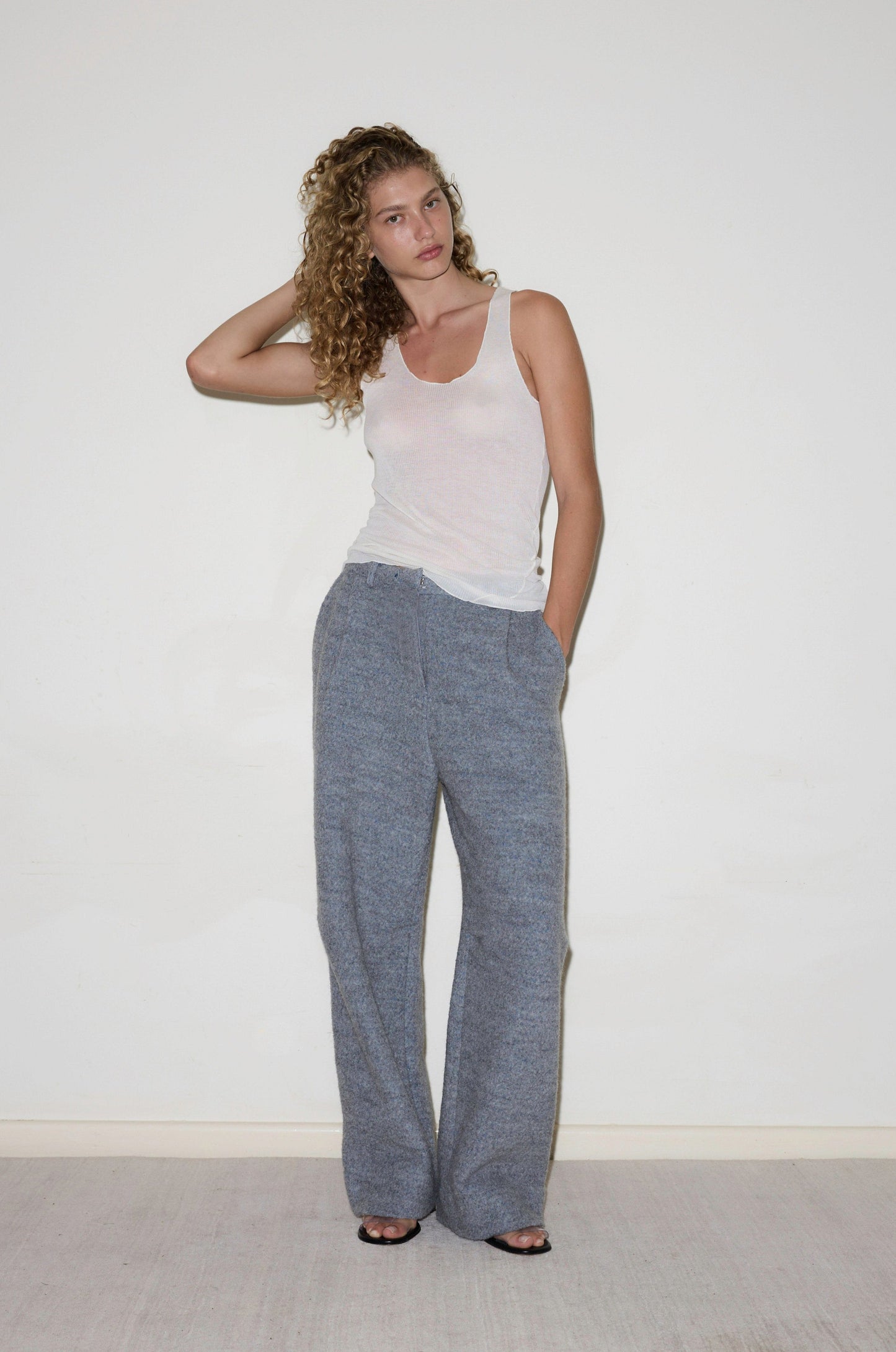 A woman with curly hair stands against a plain white wall, wearing loose gray Boiled Wool Pants, a white tank top, and black open-toed shoes. One hand is in her pocket while the other touches her hair.
