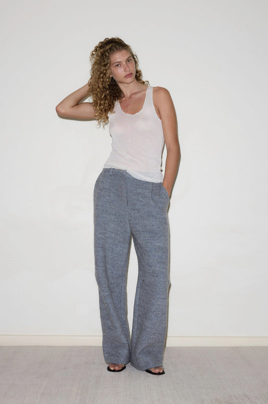 A woman with curly hair stands against a plain white wall, wearing loose gray Boiled Wool Pants, a white tank top, and black open-toed shoes. One hand is in her pocket while the other touches her hair.