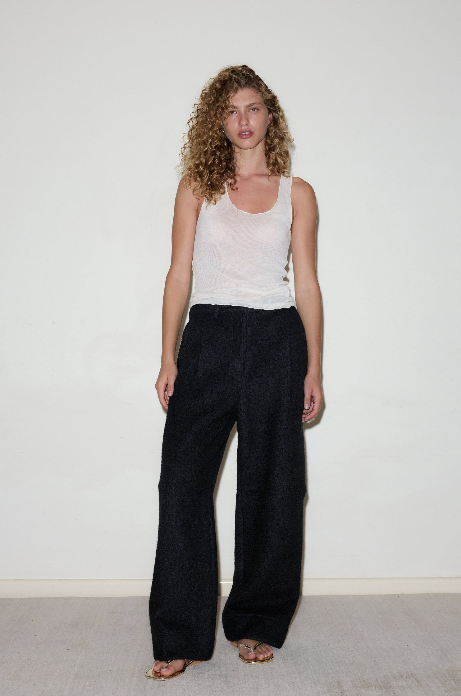 A woman with curly hair wears a sheer white tank top, sandals, and loose high-waisted Boiled Wool Pants while standing against a plain white wall on a light-colored floor.