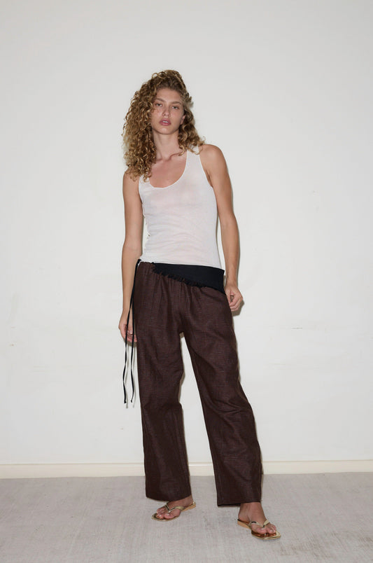 A woman with curly hair stands against a white wall, wearing a white tank top, flat sandals, and relaxed-fit Side Stripe Pant in brown tied with black strings at the waist. She poses confidently.