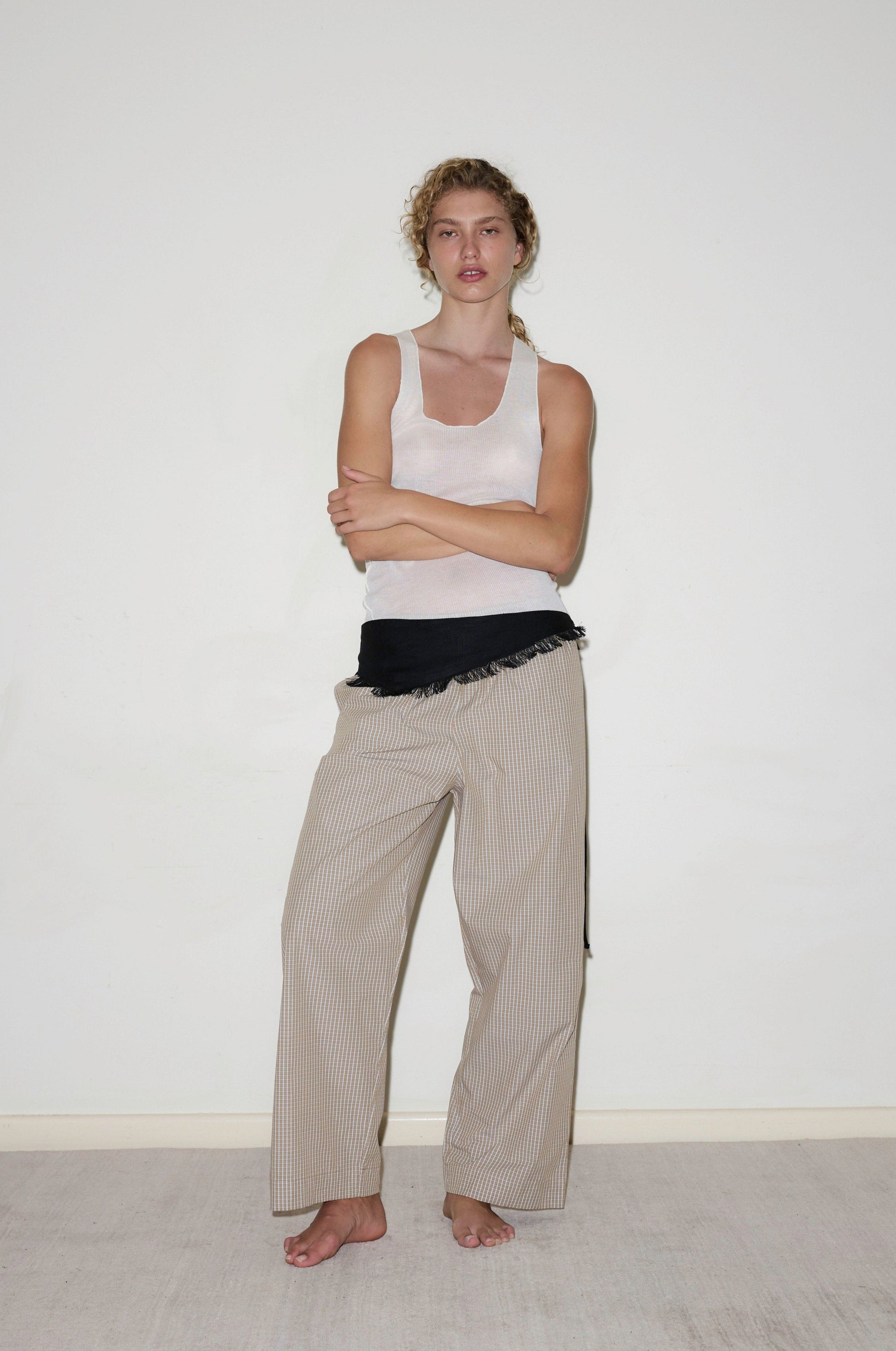 A woman with curly hair tied back stands barefoot against a plain wall, arms crossed, wearing the Deiji Studios Ease Trouser—loose, beige pinstriped pants—paired with a white tank top and a black garment tied at her waist.