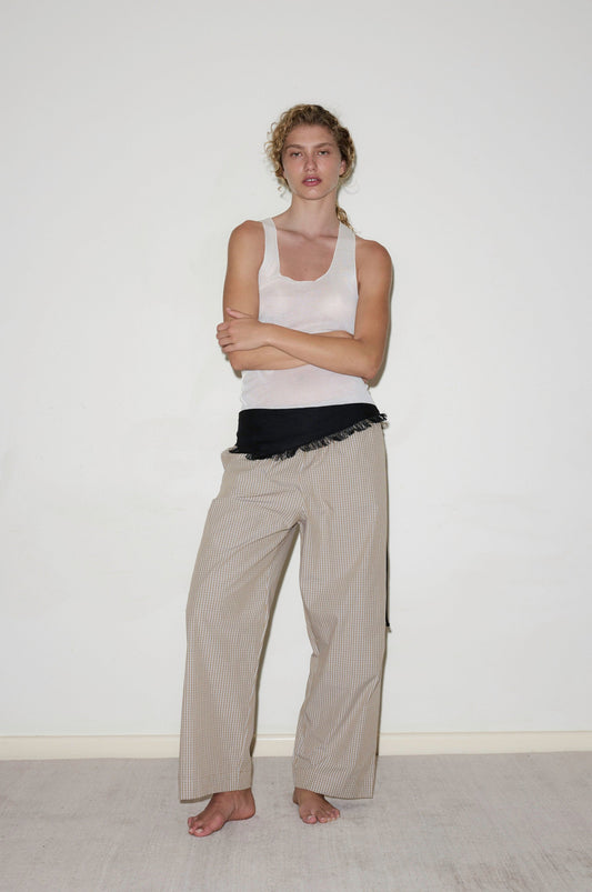 A person with curly hair stands barefoot against a white wall, arms crossed, dressed in a white tank top and the beige striped Ease Trouser, with a black garment tied around their waist.
