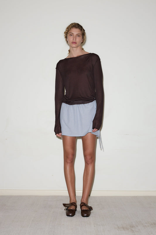 A person stands against a plain white wall wearing a long-sleeved, dark brown top, the Block Mini Skirt in blue-and-white stripes, and brown sandals for a casual, relaxed look.