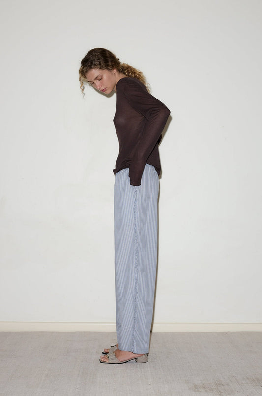 A person with curly hair stands sideways against a light-colored wall, wearing a dark long-sleeve top, Ruffle Pant in light blue stripes, and open-toe heeled sandals.