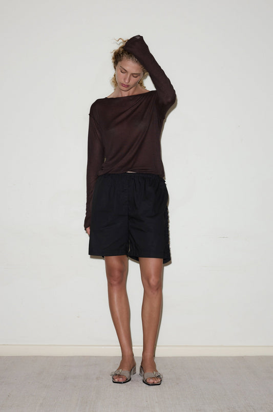 A person stands indoors against a plain white wall, wearing a long-sleeved brown top, Ruffle Short, and sandals, with one arm raised and hand resting on their head.