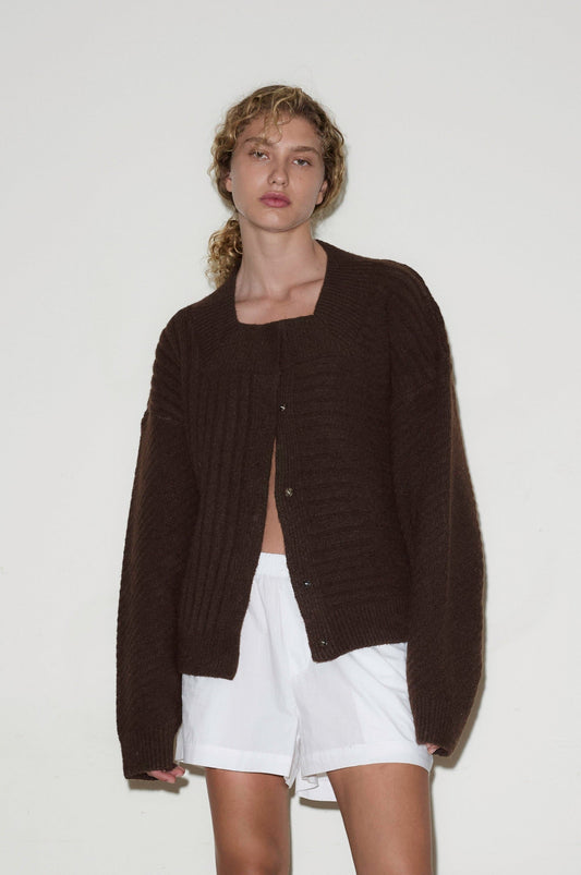 A person with curly hair wears the OS Square Cardigan in dark brown over a white top and white shorts, standing against a plain white background.