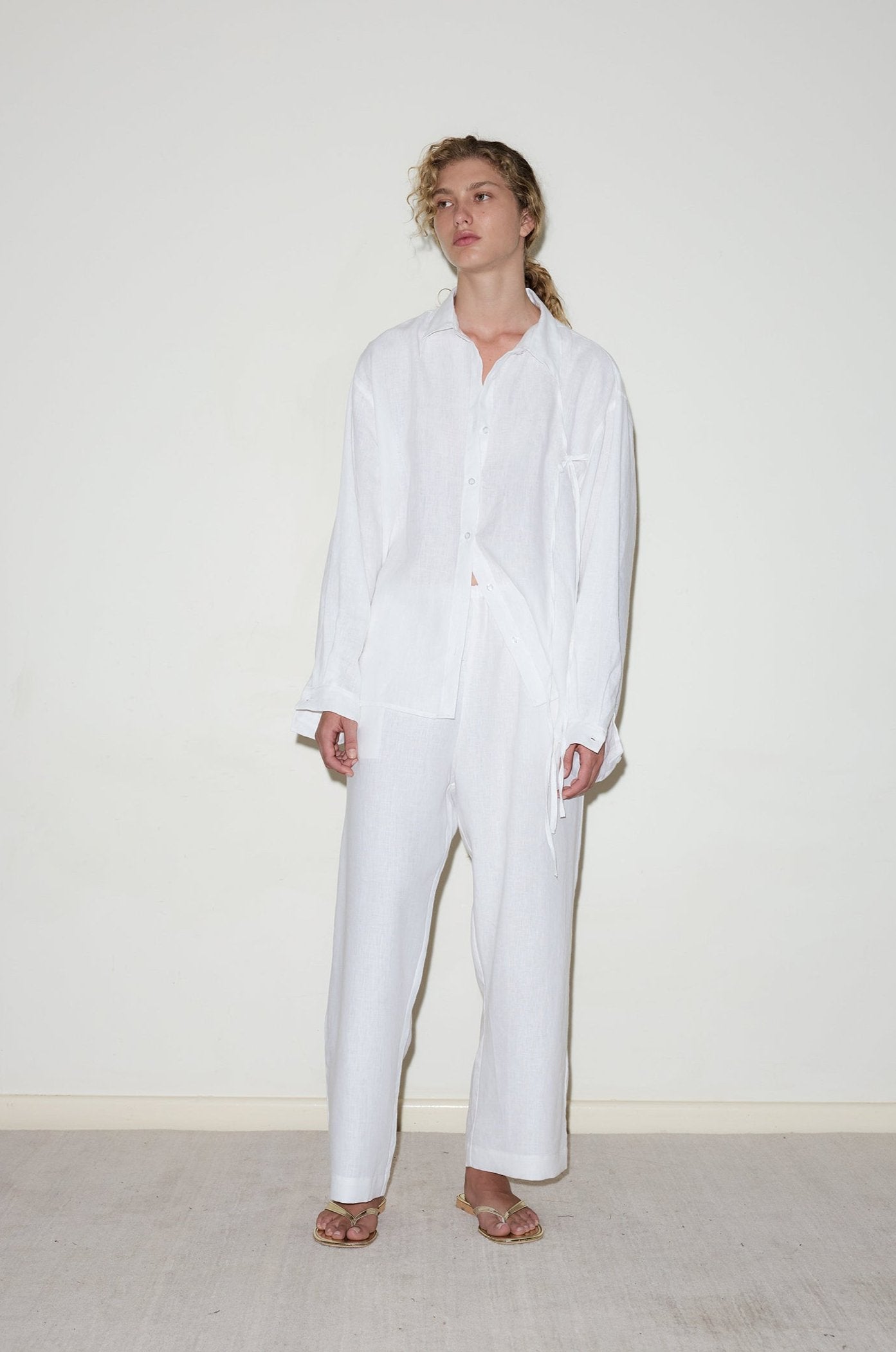 A person stands against a plain, off-white wall wearing the String Linen Set - White: a relaxed fit, stone washed French linen button-up shirt and matching pants with sandals. The minimalist ensemble looks effortlessly stylish.