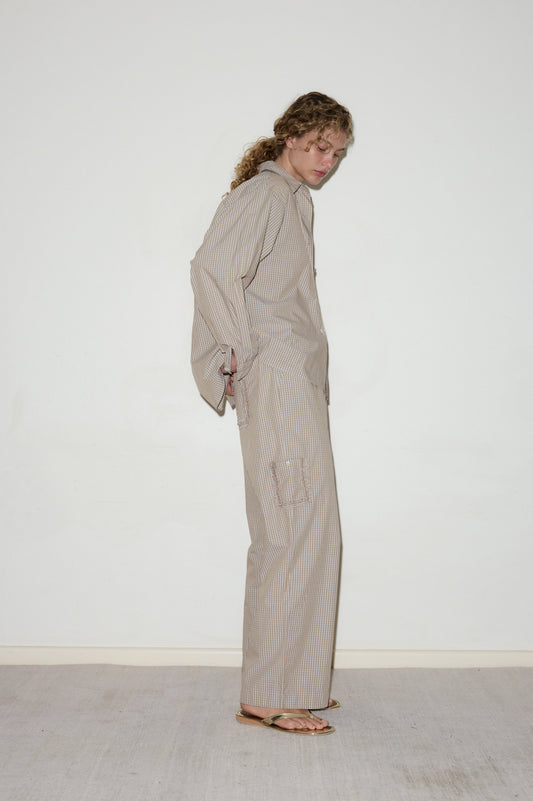 A person with curly hair stands sideways against a white wall, wearing the Cotton Pyjama Set—loose-fitting, beige and white striped long-sleeve top and matching pants with pockets—styled with brown sandals.