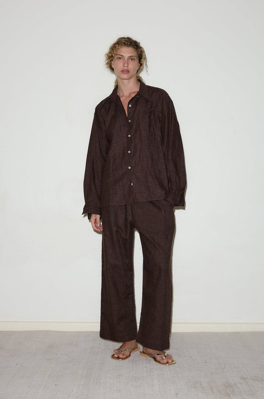 A person with curly hair stands against a plain white wall, wearing the String Linen Set—a loose, dark brown button-up shirt and matching wide-leg pants—paired with open-toed sandals.