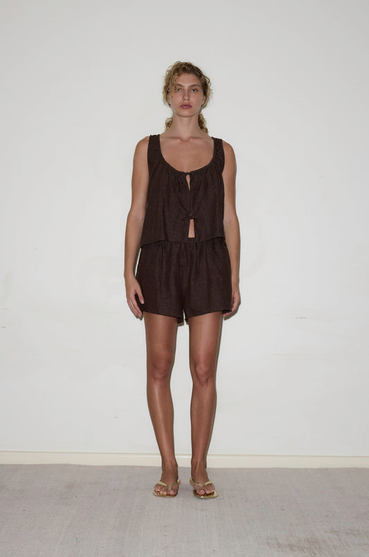 A woman stands against a white wall, wearing the Open Linen Short Set—featuring a sleeveless, loose-fitting black top with a front tie and matching shorts. Her curly hair is pulled back and she's styled with flat sandals.