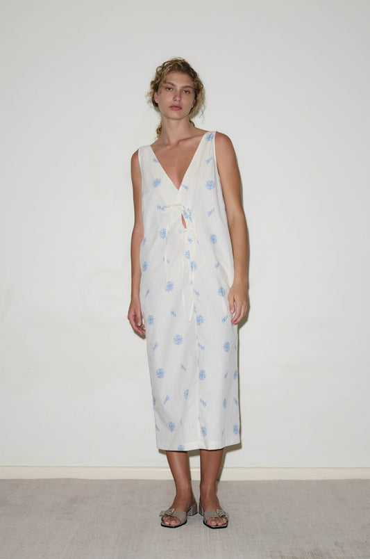 A woman with light curly hair and a relaxed posture stands against a white wall wearing the Cross Over Dress—a sleeveless white midi with blue floral prints, deep V-neck, front knot detail—and silver sandals.