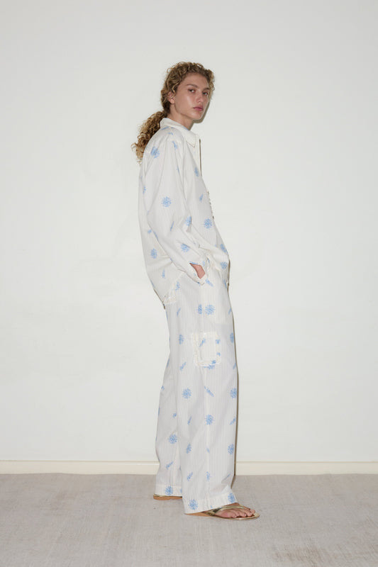 A person with curly hair stands sideways against a plain white wall, wearing the Cotton Pyjama Set—a loose-fitting white outfit with light blue patterns and sandals, hands in pockets.