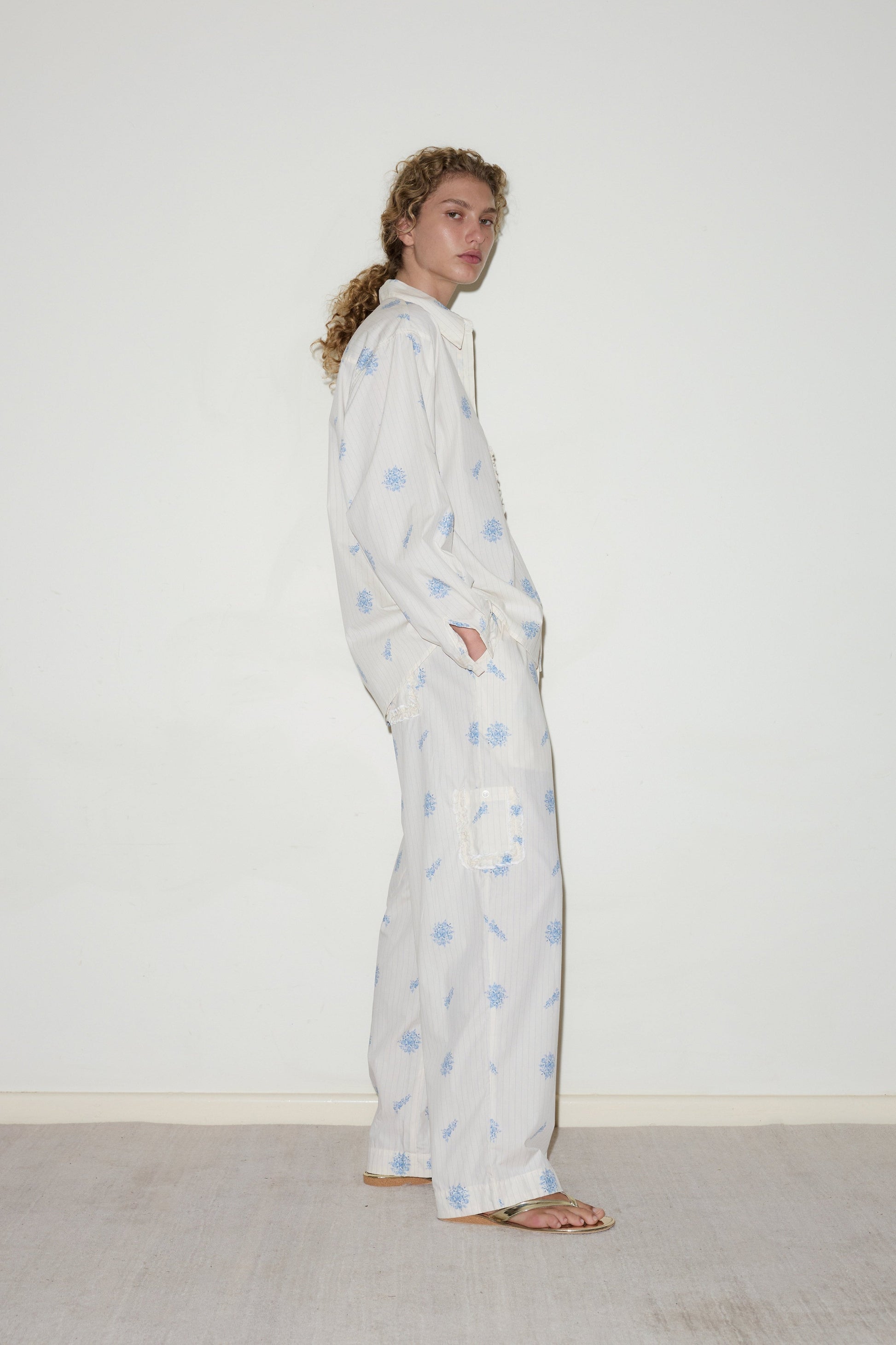A person with curly hair stands sideways against a white wall, wearing the Cotton Pyjama Set featuring loose, light fabric with blue floral patterns and sandals. Their hands are in their pockets and they look relaxed.