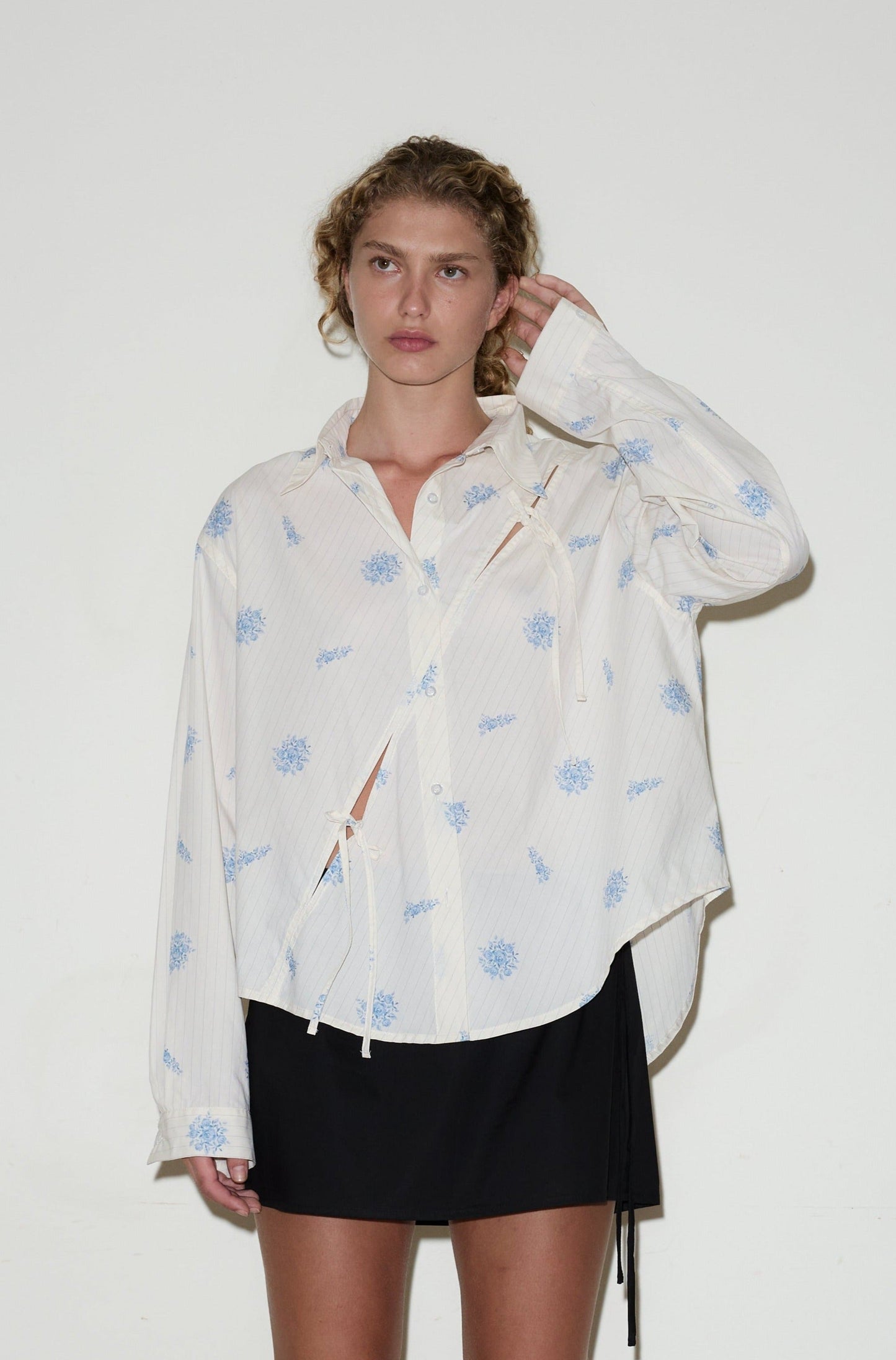 A woman with curly hair wears the twin bow shirt—a loose, white button-up with blue floral patterns—paired with a black skirt. She stands against a plain white background, one hand touching the side of her head.