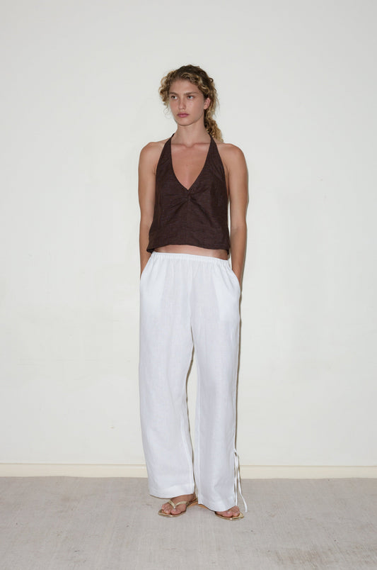 A woman stands against a plain white wall, wearing loose-fitting white Side Stripe Pants, a sleeveless dark brown halter top, and flat sandals. Her curly hair is styled loosely and her hands are in her pockets.