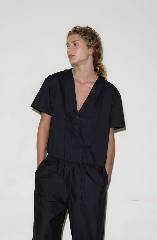 A person with curly hair stands against a plain white background, wearing the loose-fitting, short-sleeved black Panel Shirt with a V-neck and hands in their pockets, looking slightly to the side.