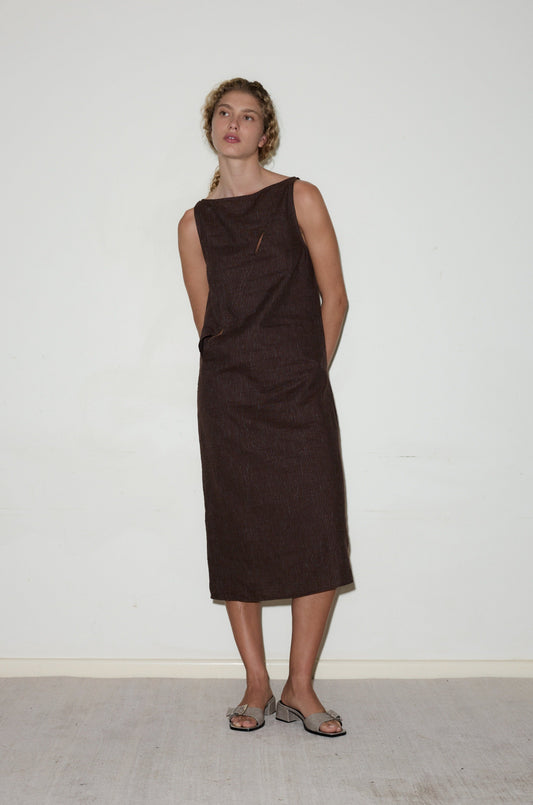 A person stands against a plain white wall, wearing the sleeveless, dark brown High Tie Dress with open-toe sandals. Their hands are behind their back, and they have curly hair styled in braids.