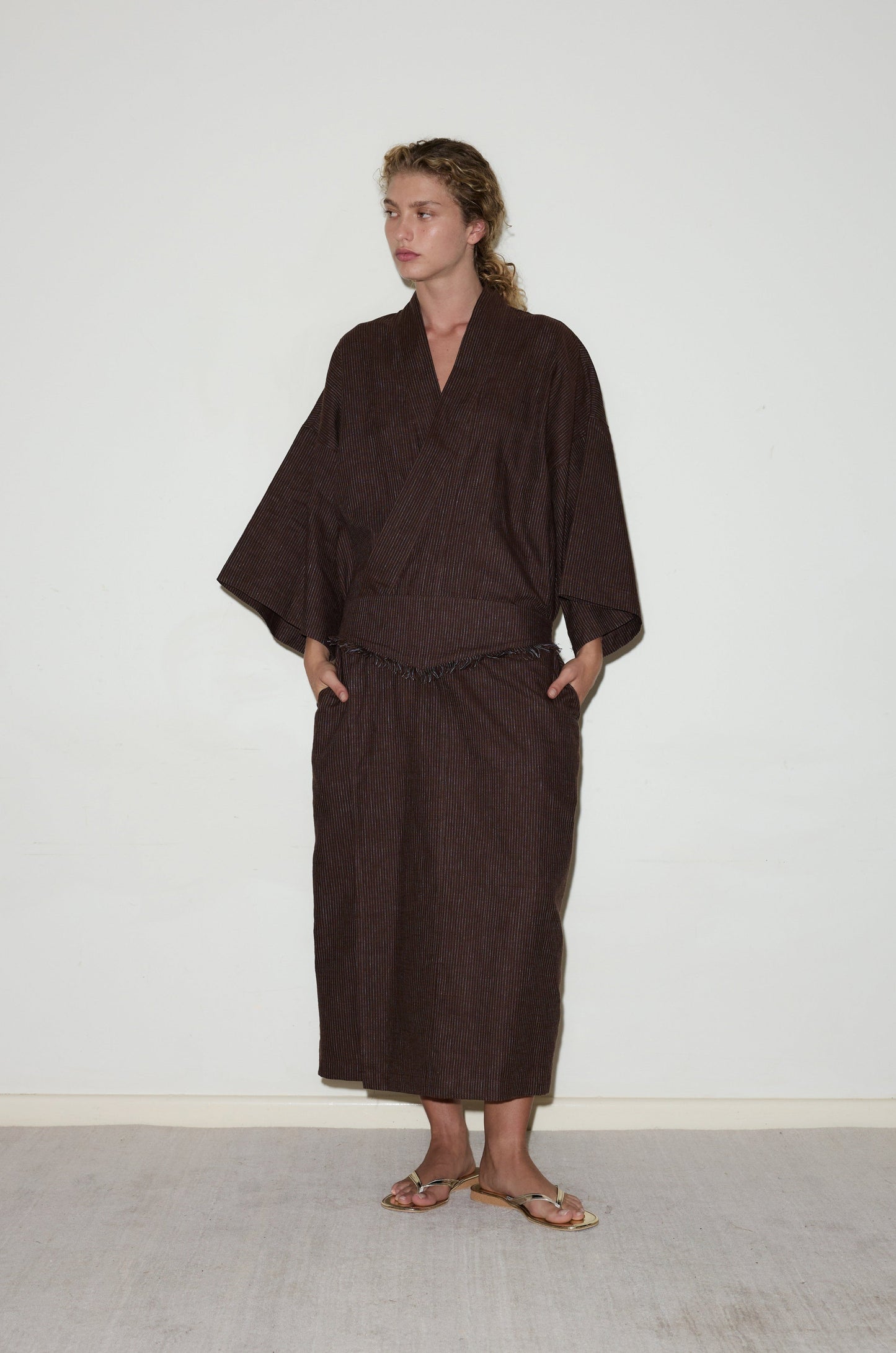 A person with light curly hair stands against a plain white wall wearing the 02 Robe, a loose dark brown kimono-style outfit with wide sleeves and a long skirt. They wear sandals and have their hands in their pockets.