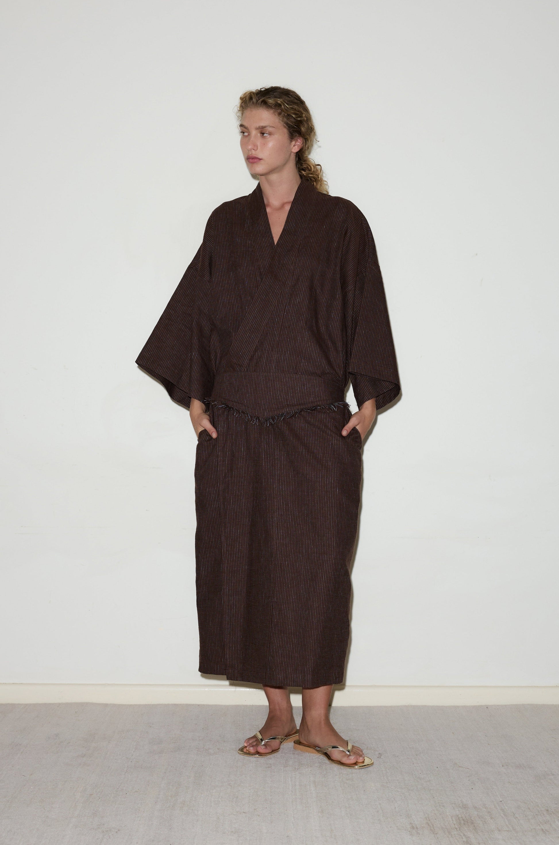 A person with light curly hair stands against a plain white wall wearing the 02 Robe, a loose dark brown kimono-style outfit with wide sleeves and a long skirt. They wear sandals and have their hands in their pockets.