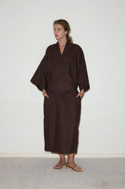 A person with light curly hair stands against a plain white wall wearing the 02 Robe, a loose dark brown kimono-style outfit with wide sleeves and a long skirt. They wear sandals and have their hands in their pockets.
