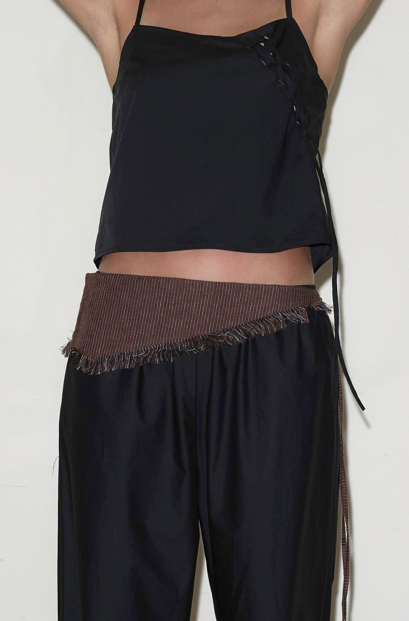 A person wears a black sleeveless top with side ties, loose black pants, and a Linen Fray Belt with brown frayed edges around their waist. Their arms are raised, and their head is out of frame.