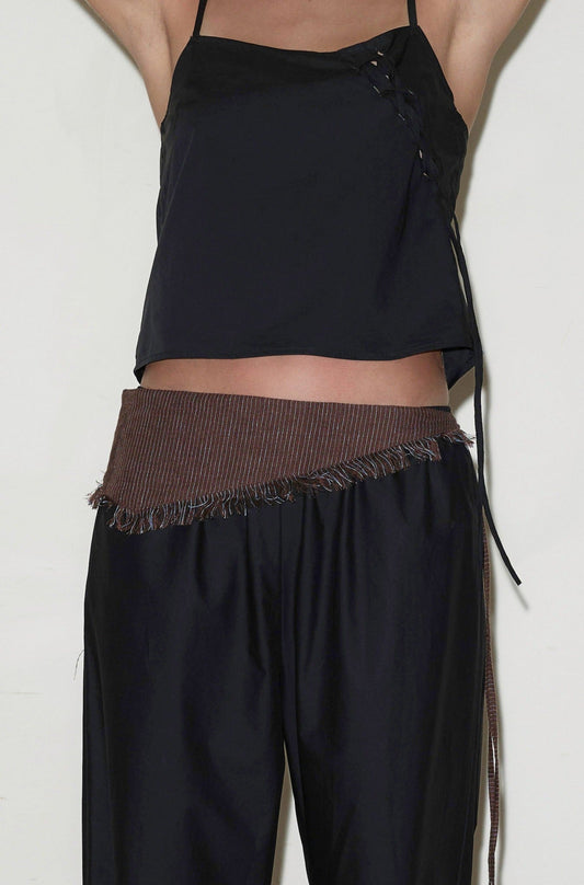 A person wears a black sleeveless top with side ties, loose black pants, and a Linen Fray Belt with brown frayed edges around their waist. Their arms are raised, and their head is out of frame.