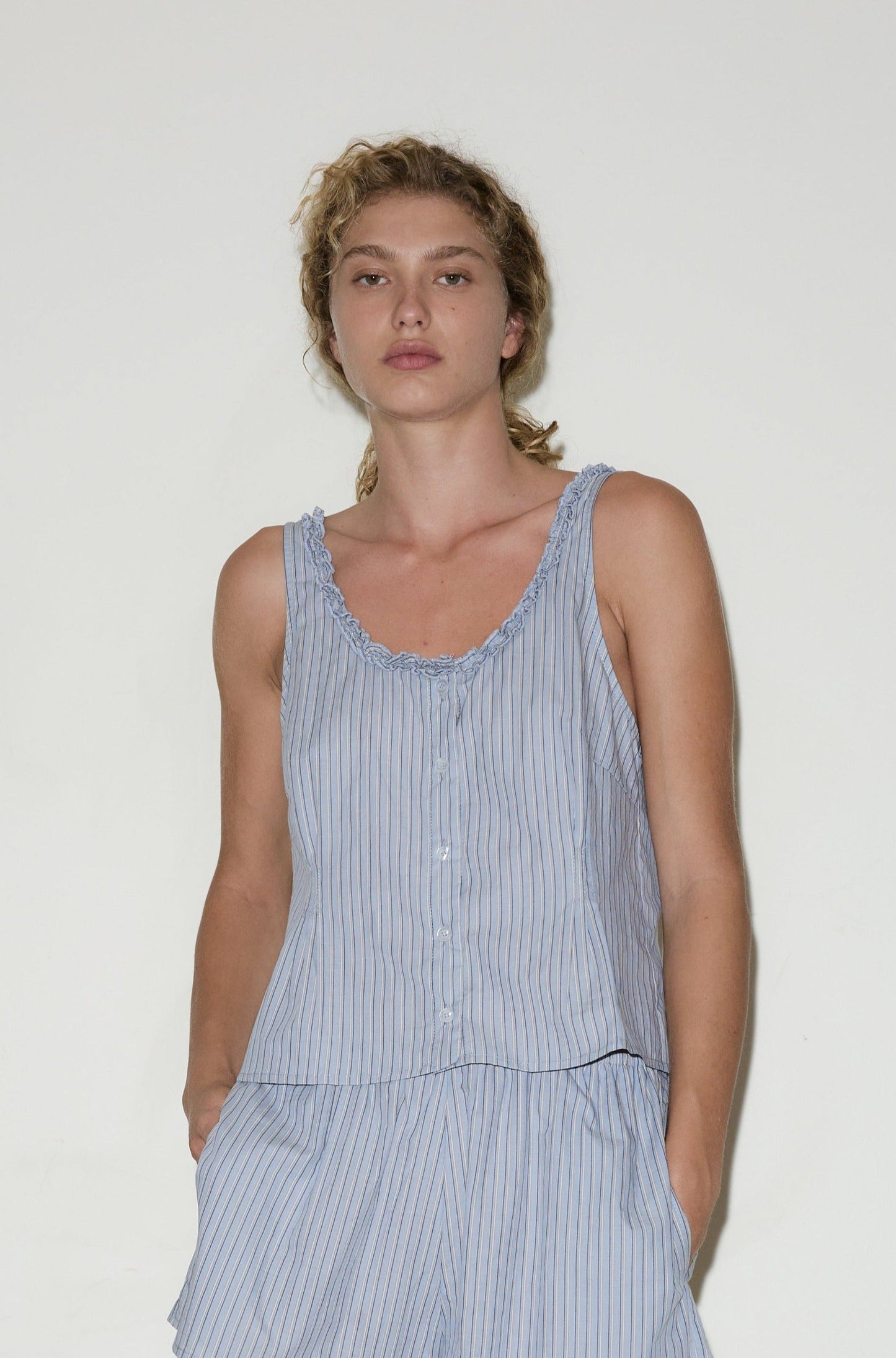 A person with blonde curly hair wears the Ruffle top, a light blue sleeveless striped top with ruffled edges, paired with matching shorts, standing against a plain white background.