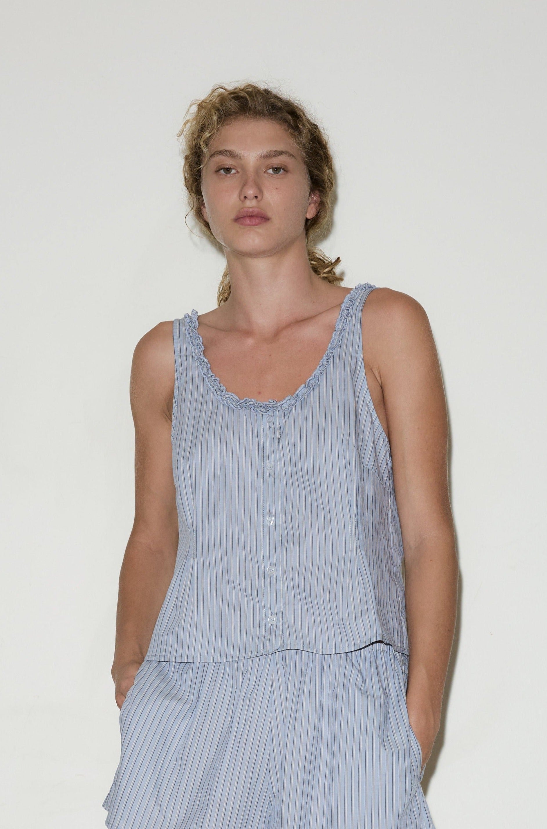 A person with blonde curly hair wears the Ruffle top, a light blue sleeveless striped top with ruffled edges, paired with matching shorts, standing against a plain white background.