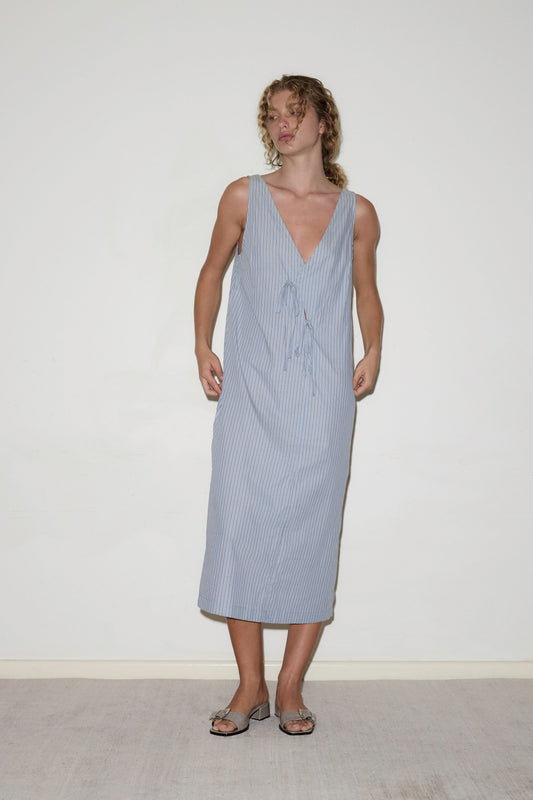 A person stands against a plain white wall, wearing the Cross Over Dress—a sleeveless, light blue and white striped midi dress with a deep V-neckline and front tie detail—paired with open-toe flat sandals.