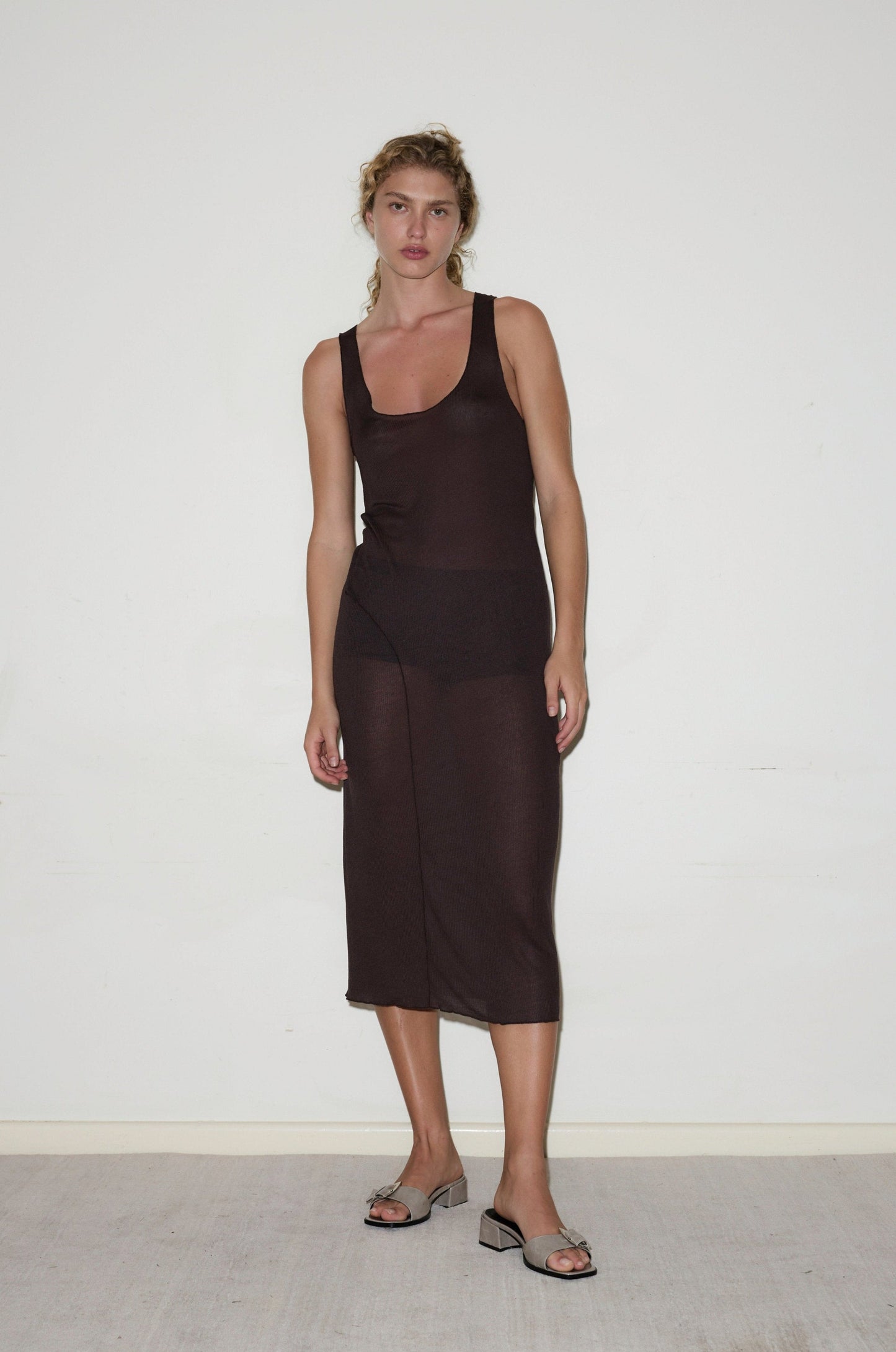 A woman with wavy hair and a relaxed expression stands against a white wall, wearing the Knit Singlet Dress - Mahogany by Deiji Studios over black undergarments, styled with open-toe heeled sandals.