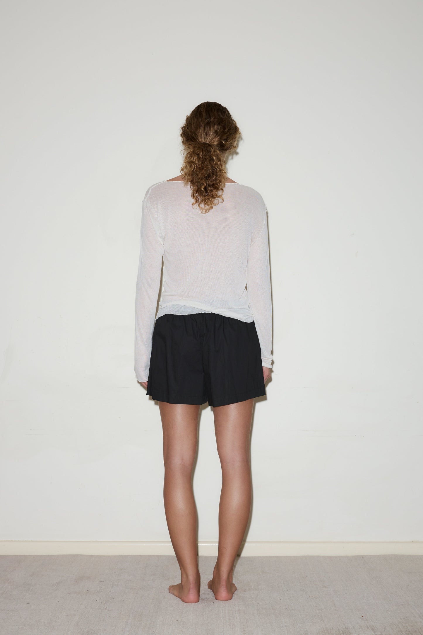 A person with curly hair stands barefoot, facing a blank white wall. They wear a long-sleeved, semi-sheer white top and the Organic Cotton Boxer - Black.