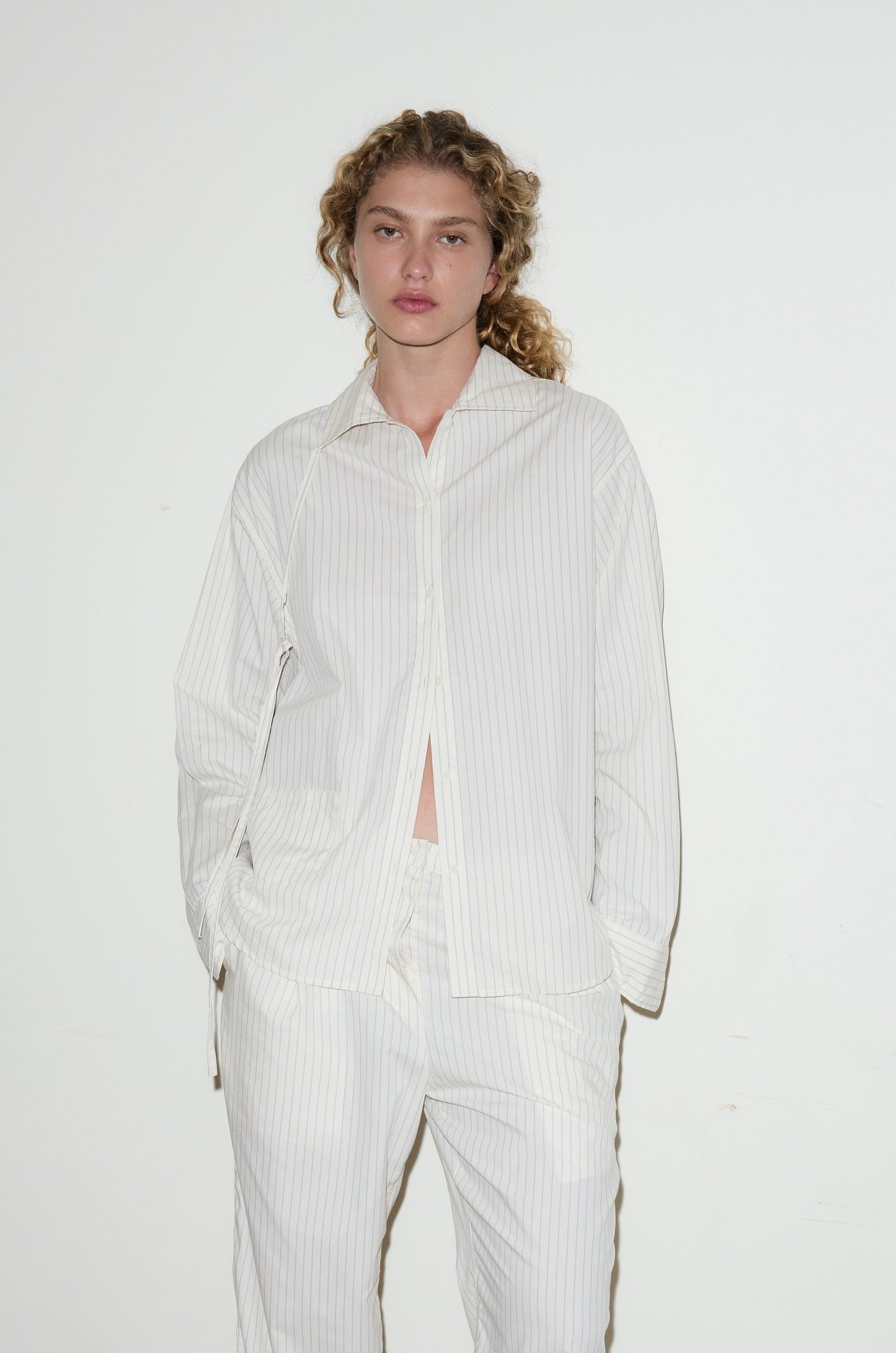 A person with curly hair stands against a white wall, wearing the Lapel Bow Shirt—a loose-fitting, white pinstripe button-up—with matching pants and hands in their pockets.